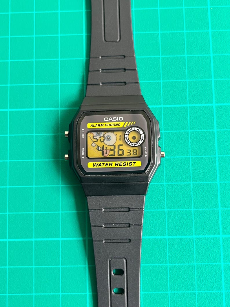 May include: Black Casio digital watch with a rectangular face and a black band. The watch face displays the time and has the words "CASIO", "ALARM CHRONO", and "WATER RESIST" printed on it. The watch is on a green surface.