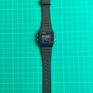 K&ouml;nnte beinhalten: Eine schwarze Digitalarmbanduhr mit rechteckigem Zifferblatt und schwarzem Armband. Das Zifferblatt zeigt Uhrzeit, Datum und weitere Informationen an. Die Uhr ist mit "CASIO F-91W" beschriftet und liegt auf einem gr&uuml;nen Rasterhintergrund.