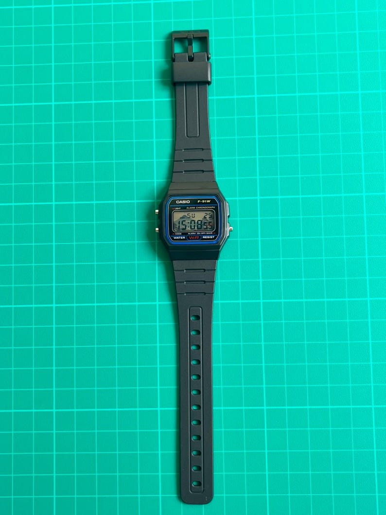 K&ouml;nnte beinhalten: Eine schwarze Digitalarmbanduhr mit rechteckigem Zifferblatt und schwarzem Armband. Das Zifferblatt zeigt Uhrzeit, Datum und weitere Informationen an. Die Uhr ist mit "CASIO F-91W" beschriftet und liegt auf einem gr&uuml;nen Rasterhintergrund.