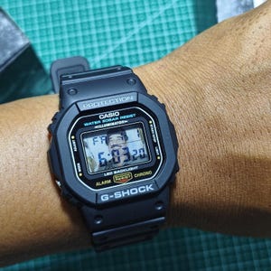 May include: A black Casio G-Shock digital watch with a rectangular face, featuring the text PROTECTION, CASIO, WATER 20BAR RESIST, ILLUMINATOR, SHOCK RESIST, CHRONO, and G-SHOCK. The digital display shows the time 6:03 and the date FR 1.