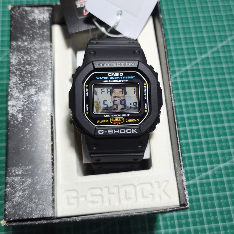 May include: A black Casio G-Shock digital watch with a square face, featuring the text PROTECTION, WATER 20BAR RESIST, ILLUMINATOR, LED BACKLIGHT, ALARM, CHRONO, and G-SHOCK. The digital display shows the time 5:59 and the date FR 1.
