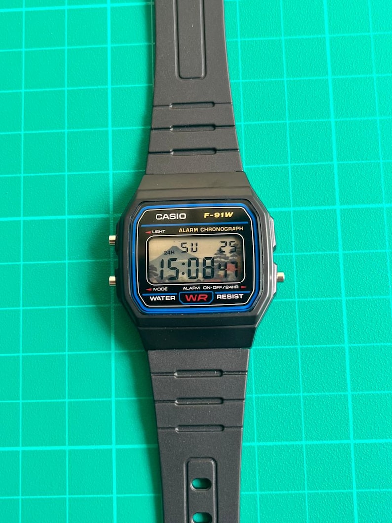 May include: Black Casio F-91W digital watch with a rectangular face and black resin strap. The watch face displays the time, day, and date digitally. Features include a light, alarm, and water resistance. The watch is approximately 3.5 cm wide.