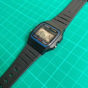 K&ouml;nnte beinhalten: Schwarze digitale Casio F-91W-Uhr mit rechteckigem Zifferblatt und schwarzem Armband. Das Zifferblatt zeigt die Uhrzeit, den Tag und das Datum an. Die Uhr hat blaue und silberne Akzente und der Text "CASIO" und "F-91W" sind sichtbar.