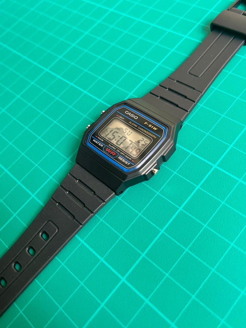 K&ouml;nnte beinhalten: Schwarze digitale Casio F-91W-Uhr mit rechteckigem Zifferblatt und schwarzem Armband. Das Zifferblatt zeigt die Uhrzeit, den Tag und das Datum an. Die Uhr hat blaue und silberne Akzente und der Text "CASIO" und "F-91W" sind sichtbar.
