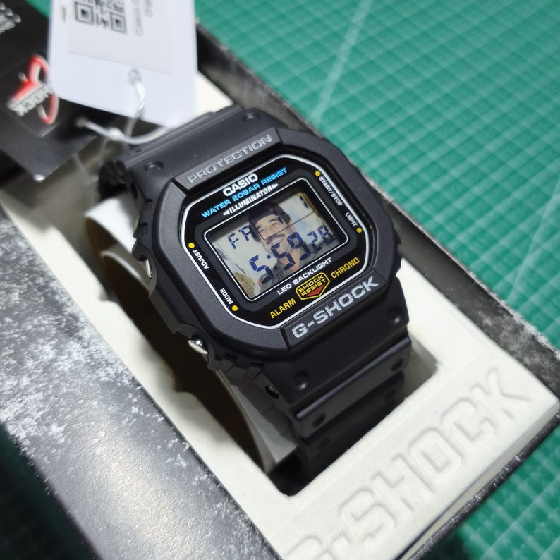 May include: A black Casio G-Shock digital watch with a rectangular face, featuring the text PROTECTION, WATER 20BAR RESIST, ILLUMINATOR, SHOCK RESIST, and CHRONO. The digital display shows the time 5:59 and the day FR, housed in its original packaging.