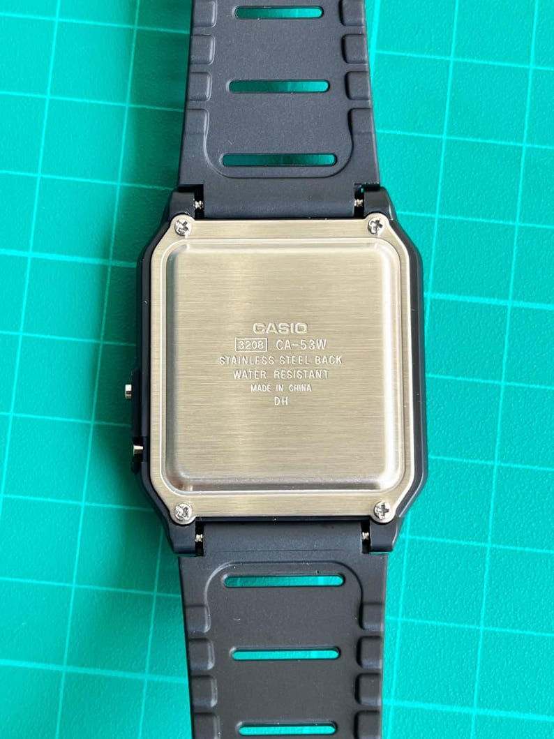 May include: Close-up of a Casio CA-53W digital watch. The watch has a black resin strap and a stainless steel back. The back of the watch has the Casio logo and model information, including "WATER RESISTANT" and "MADE IN CHINA".