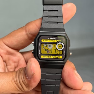 May include: Black Casio digital watch with a yellow face featuring a cartoon character. The watch has a black band and the text "CASIO", "ALARM CHRONO", and "WATER RESIST" are visible. The watch is held by a hand.