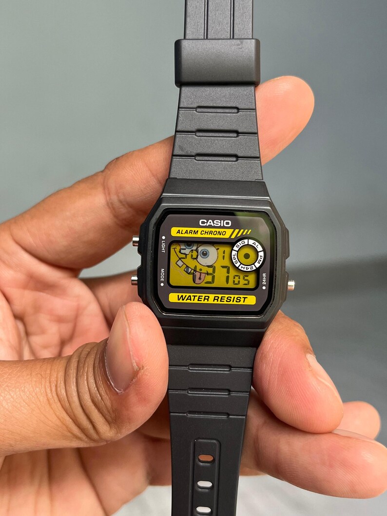 May include: Black Casio digital watch with a yellow face featuring a cartoon character. The watch has a black band and the text "CASIO", "ALARM CHRONO", and "WATER RESIST" are visible. The watch is held by a hand.