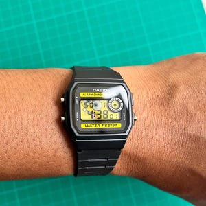May include: Black Casio digital watch with a rectangular face and a black band. The watch face displays the time and has the words "CASIO", "ALARM CHRONO", and "WATER RESIST". The watch is on a wrist.
