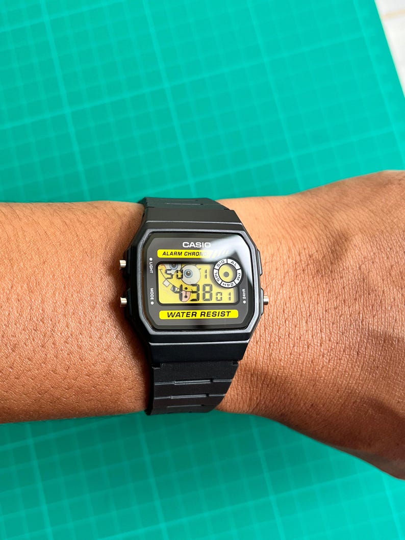 May include: Black Casio digital watch with a rectangular face and a black band. The watch face displays the time and has the words "CASIO", "ALARM CHRONO", and "WATER RESIST". The watch is on a wrist.