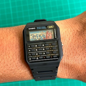 May include: Black Casio digital watch with calculator buttons and a rectangular face. The watch displays the time and has the text "CASIO WATER RESIST ALARM CHRONO WR" on the face. The watch is worn on a wrist.