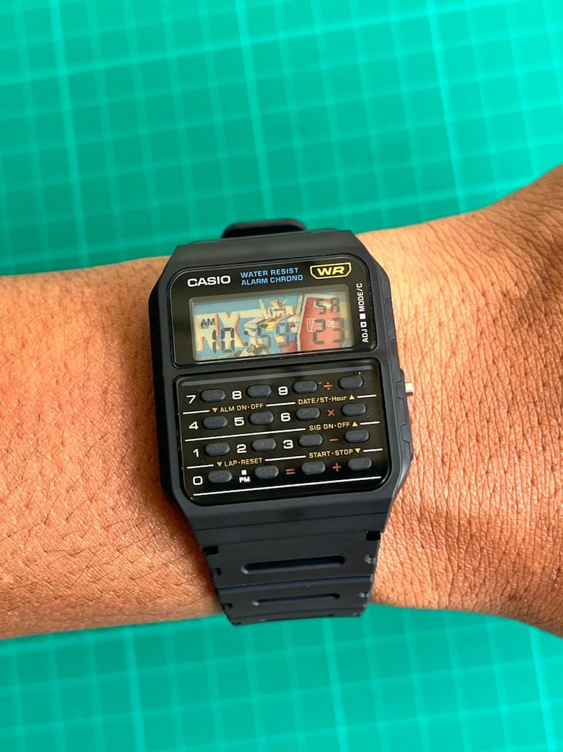 May include: Black Casio digital watch with calculator buttons and a rectangular face. The watch displays the time and has the text "CASIO WATER RESIST ALARM CHRONO WR" on the face. The watch is worn on a wrist.