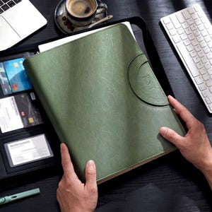 Peut inclure: Un porte-documents en cuir vert sauge avec une fermeture éclair et un rabat arrondi. Le rabat est estampé des initiales "R.S.T.". Le porte-documents est ouvert, révélant des fentes pour cartes et un stylo. Un ordinateur portable et un clavier sont en arrière-plan.