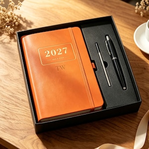 May include: An orange notebook with the year 2027 and the initials TW embossed on the cover, presented in a black gift box alongside a black ballpoint pen and a thin metal stylus. The notebook features a vertical elastic band closure.