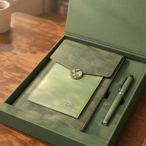 May include: A green stationery set in a presentation box. The set includes a notebook, a pen, and a small pouch with a decorative button. The notebook has the initials "BE" embossed on the front.