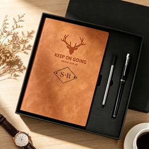 May include: A brown leather journal with a deer head logo and the text "KEEP ON GOING NEVER GIVE UP S.R." is presented in a black gift box, accompanied by a silver pen and a black pen. A watch and coffee cup are also visible.