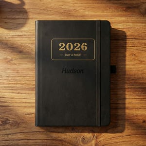 Peut inclure: Agenda noir 2026, un jour par page, avec une étiquette rectangulaire dorée. L'étiquette affiche l'année et les mots "DAY A PAGE". Le nom "Hudson" est imprimé en dessous. Le journal a une fermeture élastique.