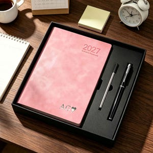 May include: A pink 2027 schedule notebook with a textured cover, featuring the silver embossed letters AG and a heart symbol. The notebook is presented in a black gift box alongside a black ballpoint pen and a thin silver metal stylus.