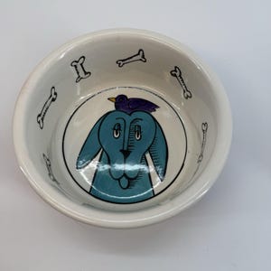 May include: White ceramic pet bowl with a blue dog illustration and a purple bird on its head. The bowl has black bone designs around the rim. A fun and decorative bowl for your pet.