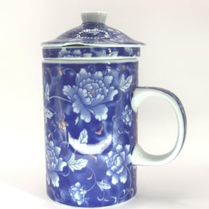 May include: A blue and white ceramic tea mug with a lid, featuring a floral peony design. The mug has a handle and a matching lid with a small knob. The design includes light blue flowers and green leaves on a dark blue background.