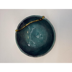 May include: A handcrafted teal ceramic bowl with a textured surface. A gold-colored accent piece extends across the rim. The bowl has a rounded shape and is suitable for serving or display.