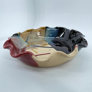 May include: A handcrafted ceramic bowl with a unique, ruffled edge. The bowl features a color-blocked design with sections of red, beige, black, blue, and brown. The interior has a glossy finish, and the bowl is ideal for serving or display.