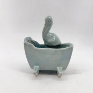 May include: A light blue ceramic planter shaped like a small bathtub with a sculpted pelican figure attached to the back rim. The piece features a smooth, glossy finish with subtle speckled details and stands on four short, sturdy legs.