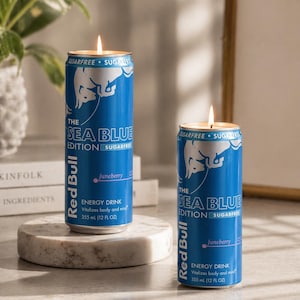 May include: Two blue Red Bull energy drink cans repurposed as candles. The cans are labeled "The Sea Blue Edition" and "Juneberry". Each candle has a lit wick, and the cans are on a marble surface.