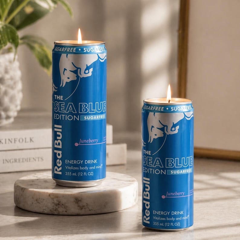 May include: Two blue Red Bull energy drink cans repurposed as candles. The cans are labeled "The Sea Blue Edition" and "Juneberry". Each candle has a lit wick, and the cans are on a marble surface.