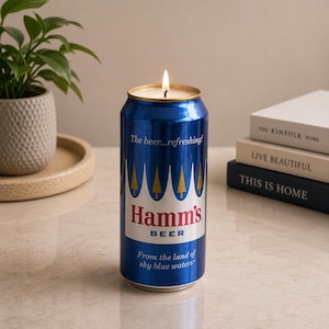 May include: A blue Hamm's beer can candle with a lit wick. The can has the Hamm's Beer logo and the text "From the land of sky blue waters." The candle sits on a light-colored surface with a plant and books in the background.