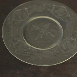 May include: A clear glass plate with an etched floral design. The plate has a circular shape with a slightly raised rim. The etched design includes flowers and leaves around the edge and in the center.