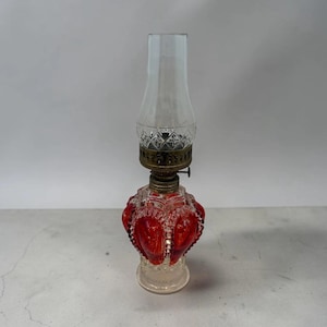 May include: A vintage oil lamp with a clear glass chimney and a ruby red glass base. The base has a decorative, heart-shaped design. The lamp has a brass-colored metal fitting and a clear glass base.