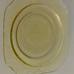 May include: A vintage yellow glass plate with a square shape and rounded corners. The plate features an embossed floral pattern around the edges and a central diamond design. The plate is approximately 9 inches in diameter.