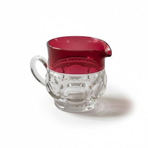 May include: A glass creamer pitcher featuring a clear, textured base with a repeating circular pattern and a smooth, deep red rim. The pitcher includes a curved handle on the side and a small pouring spout, showcasing a two-tone design.