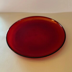 May include: A round, red glass plate with a black rim. The plate has a glossy finish and is set against a neutral background. The plate is a solid color and appears to be new.