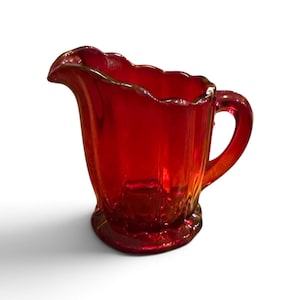 May include: A vibrant ruby red glass pitcher featuring a scalloped rim, a curved handle, and a fluted body design. The translucent vessel stands on a circular base with a matching scalloped edge, showcasing a deep crimson hue throughout the glass.