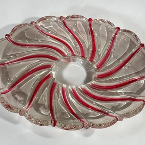 May include: A clear glass bowl with a circular center opening and a scalloped edge. The bowl features a radial pattern of swirling, opaque red glass stripes that extend from the center toward the outer rim, creating a distinct spiral effect.