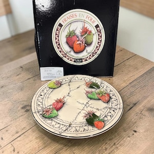 May include: A round ceramic plate featuring a cream-colored base with black line-drawn patterns and raised, three-dimensional strawberry motifs. The plate is displayed in front of a black box labeled Fraises en Folie with a matching strawberry graphic.