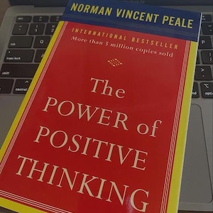 Può includere: Un libro intitolato "The Power of Positive Thinking" di Norman Vincent Peale. La copertina rossa presenta testo bianco e un bordo giallo. Il libro è su una tastiera di un laptop.