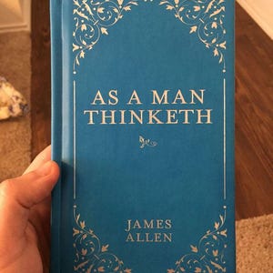 May include: A teal-colored book titled "AS A MAN THINKETH" by James Allen. The book has decorative gold floral corner designs and a red bookmark. The book is held by a person.
