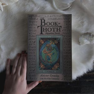 Puede incluir: El Libro de Thoth (Tarot Egipcio) de Aleister Crowley. La portada del libro presenta una ilustraci&oacute;n de una figura entrelazada con una serpiente, rodeada de dise&ntilde;os ornamentados y jerogl&iacute;ficos. El libro est&aacute; sobre una superficie blanca similar a la piel.