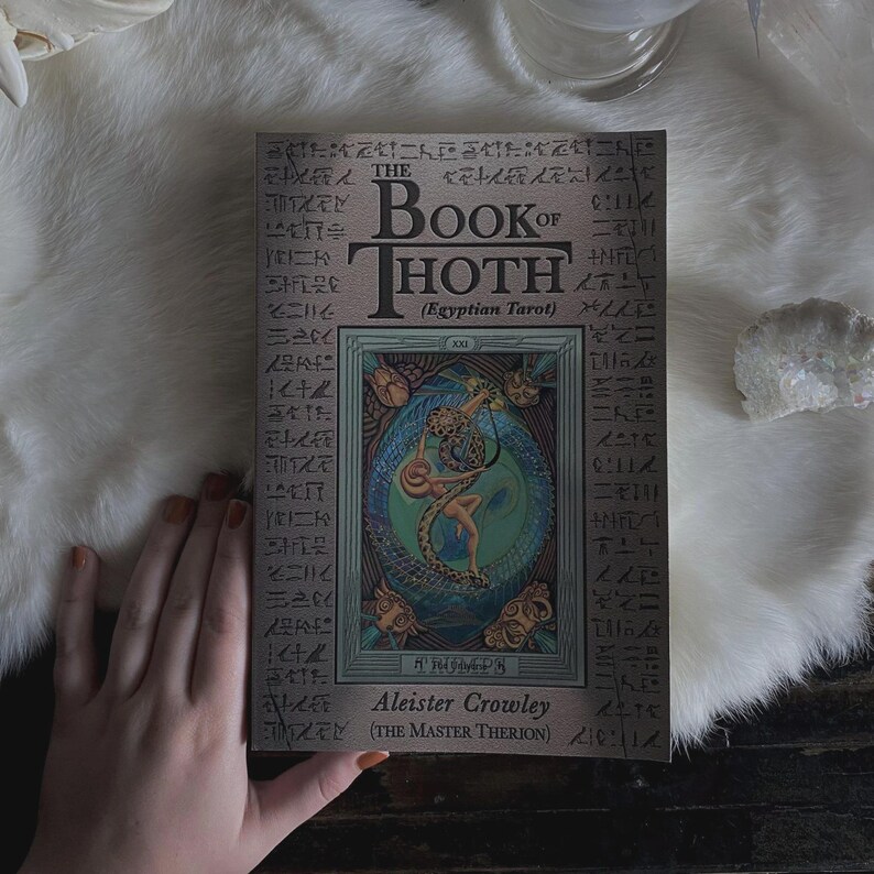 Puede incluir: El Libro de Thoth (Tarot Egipcio) de Aleister Crowley. La portada del libro presenta una ilustraci&oacute;n de una figura entrelazada con una serpiente, rodeada de dise&ntilde;os ornamentados y jerogl&iacute;ficos. El libro est&aacute; sobre una superficie blanca similar a la piel.