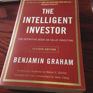 Op de afbeelding: Een rood boek getiteld "The Intelligent Investor" van Benjamin Graham. Het boek heeft een gouden rand en de tekst "Revised Edition". De omslag bevat een citaat van Warren Buffett. Het boek gaat over value investing.