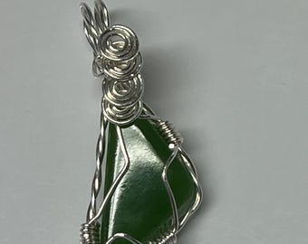 May include: A handcrafted pendant featuring a polished, emerald-green stone encased in intricate silver wire. The wirework includes decorative swirls at the top and wraps around the stone's edges, creating a unique, artistic piece of jewelry.