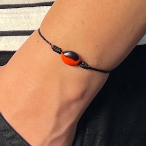 May include: Black string bracelet with a central bead. The bead is split, with a red half and a black half. The bracelet is knotted and worn on a wrist, with a black pocket and striped shirt in the background.