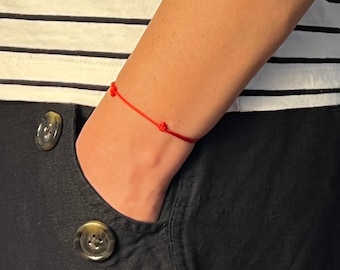 May include: A red string bracelet with three knots is worn on a person's wrist. The person is wearing a black and white striped shirt and black pants. The bracelet is a simple, minimalist design.