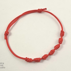 May include: A red string bracelet with multiple knots. The bracelet is made of a thin, flexible cord and features several evenly spaced knots along its length. The bracelet is designed to be adjustable and tied to fit the wrist.
