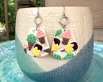 May include: A pair of whimsical earrings featuring a white base adorned with colorful ice cream cone and popsicle designs. The earrings have silver-tone ear wires and are displayed on a textured ceramic vase.