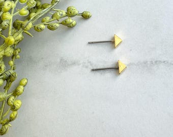 May include: A pair of gold-colored triangle stud earrings with silver posts. The earrings are displayed on a white and gray marble surface, with a sprig of green and yellow plant life in the upper left corner.