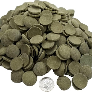 May include: A large pile of olive-green, circular, flat tablets. The tablets are stacked and scattered, with a U.S. dime for size comparison. The tablets have a slightly rough texture and appear to be a food product.